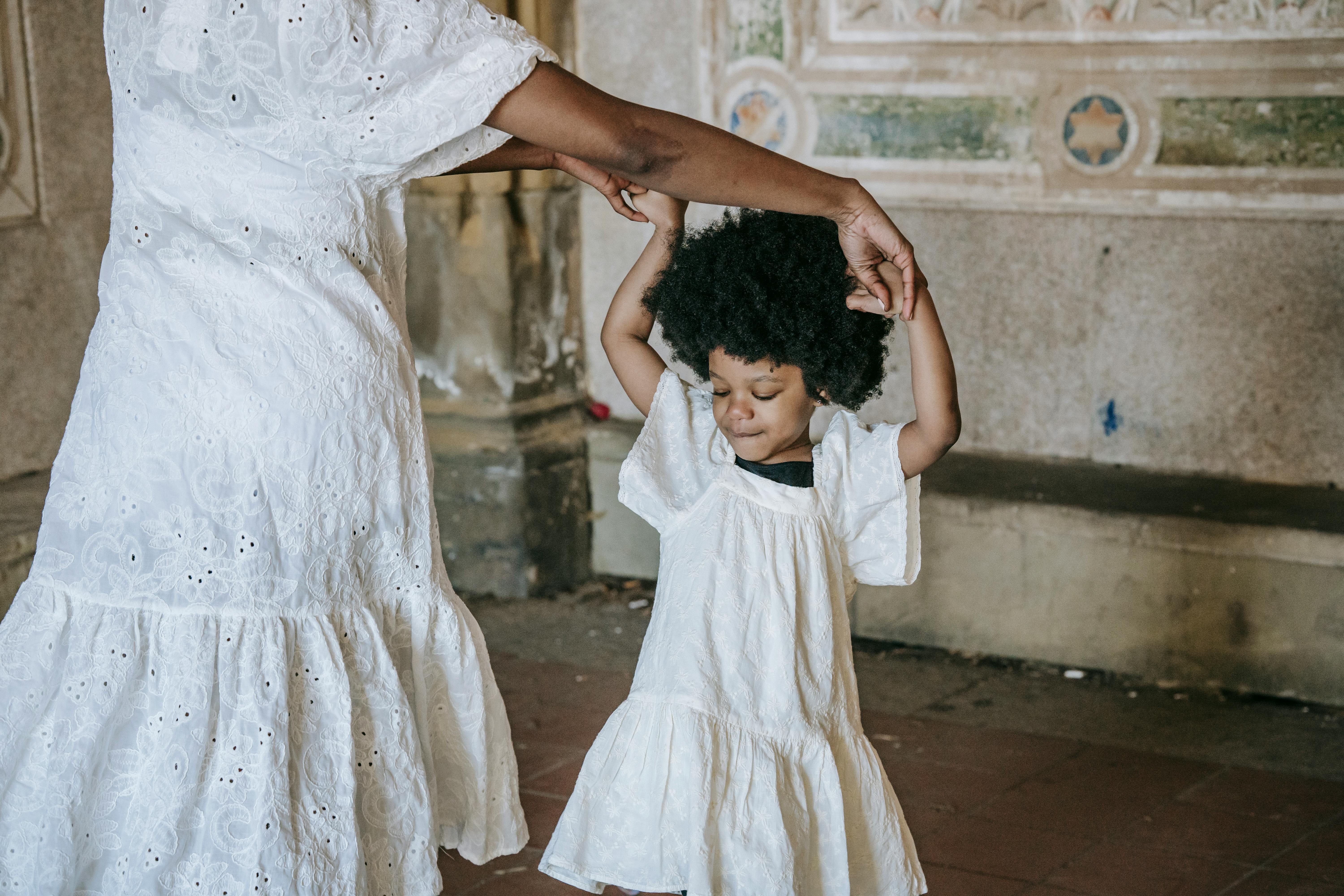 A tender moment as a mother dances with her daughter in matching white dresses indoors.